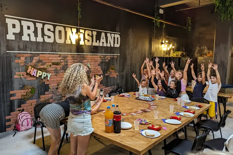 Crianças à mesa levantam os braços diante de um bolo, por baixo do letreiro da Prison Island